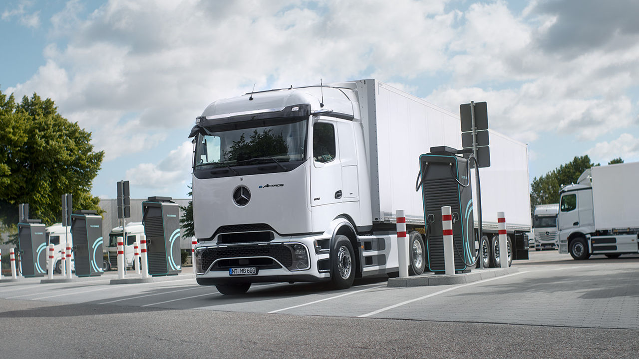 Een witte eActros 600 met ProCabin van Mercedes-Benz Trucks met een witte oplegger staat op een parkeerplaats en wordt bij een laadstation opgeladen. Rond de vrachtwagen zijn meerdere andere laadstations en afzetpalen zichtbaar. Op de achtergrond staan nog meer witte vrachtwagens, en aan de linkerkant van het beeld zijn groene bomen te zien onder een gedeeltelijk bewolkte hemel.