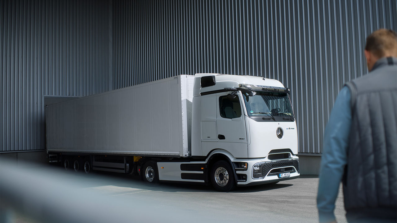 Een witte eActros 600 met ProCabin van Mercedes-Benz Trucks en een lange, witte oplegger staat op een geasfalteerd terrein voor een grijze golfplatenwand. De vrachtwagen is van opzij zichtbaar, waarbij de voorkant iets naar links wijst. Op de voorgrond staat, met de rug naar de kijker, een persoon in een blauwe jas en een grijze bodywarmer die naar de vrachtwagen kijkt. Het licht valt van boven op de scène.