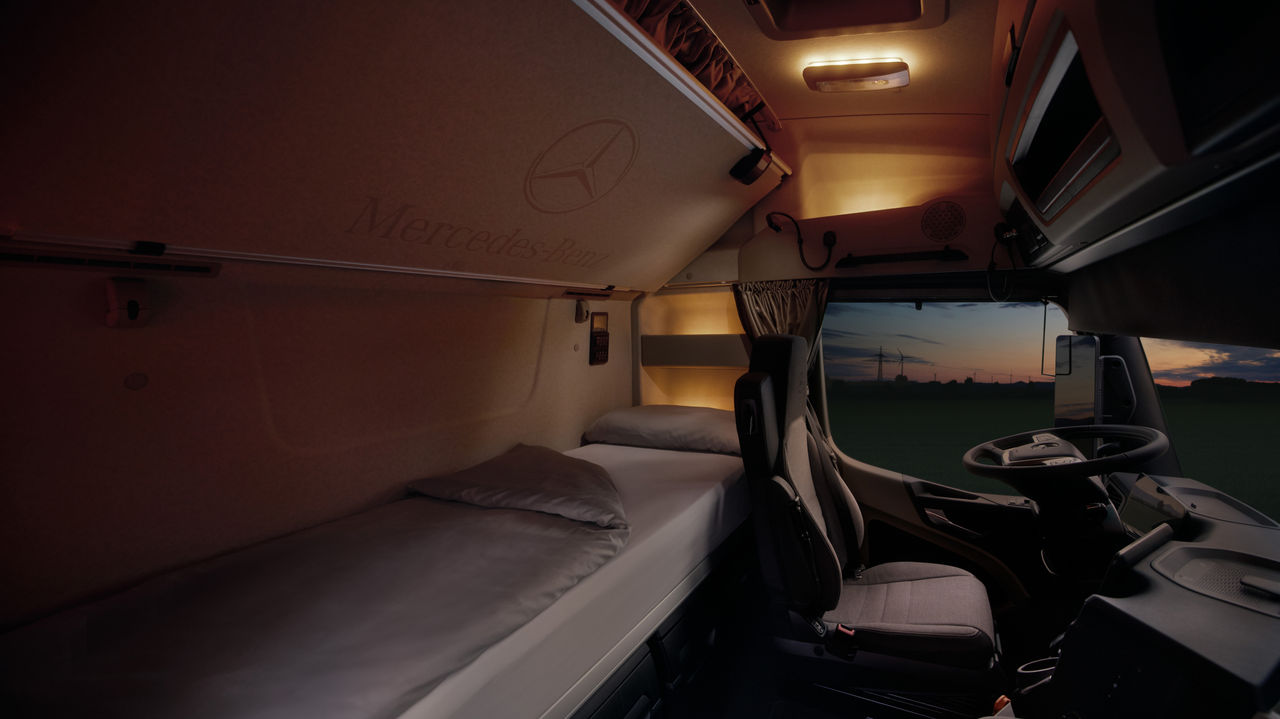 De afbeelding toont het interieur van de cabine van een eActros van Mercedes‑Benz Trucks bij schemering. Links is een keurig opgemaakt bed met lichtgrijs beddengoed te zien; daarboven aan het plafond het Mercedes‑Benz-logo en de tekst 'Mercedes‑Benz'. Rechts daarvan bevinden zich de bestuurdersstoel en het stuur, terwijl door de voorruit een donkere hemel met de silhouetten van windturbines en een uitgestrekt landschap zichtbaar is.
