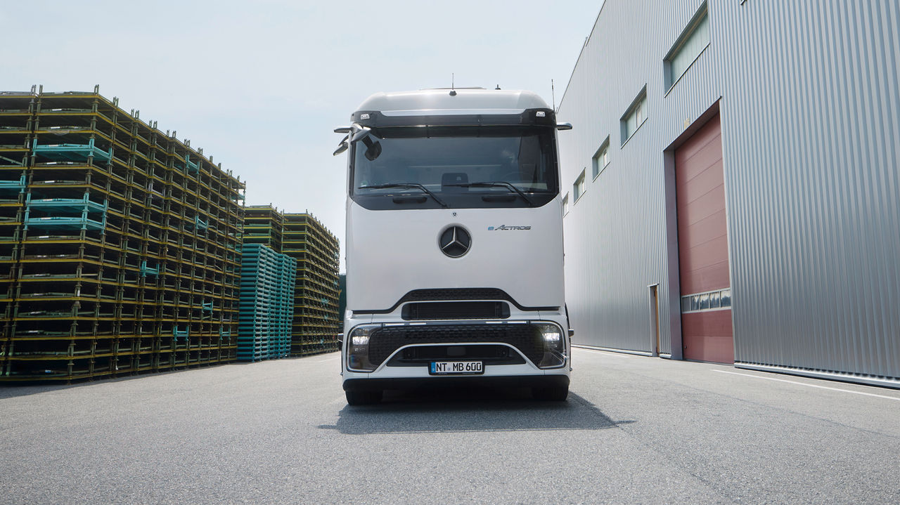 Een eActros 600 met ProCabin van Mercedes-Benz Trucks staat frontaal op een geasfalteerd terrein. Links in beeld zijn hoge stapels gele en blauwe pallets te zien, terwijl rechts een groot, modern gebouw met een grijze golfplatengevel en een rode poort staat. De lucht is helder en licht bewolkt.