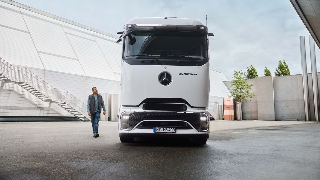 Een man in werkkledij loopt over een geasfalteerde oppervlakte, terwijl op de achtergrond een witte eActros 400 met ProCabin van Mercedes-Benz Trucks geparkeerd staat. Links is een modern gebouw met een lange buitentrap te zien en rechts bomen en andere gebouwstructuren.