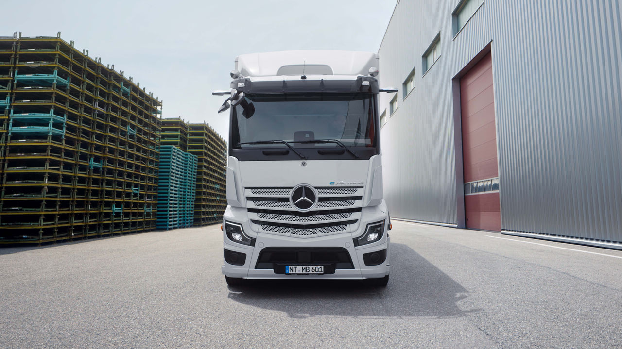 Een eActros 600 van Mercedes‑Benz Trucks staat frontaal op een geasfalteerd terrein voor een modern gebouw met een lange buitentrap. De vrachtwagen is wit en heeft het Mercedes‑sterlogo op de grille, terwijl op de achtergrond bomen en andere gebouwen zichtbaar zijn.