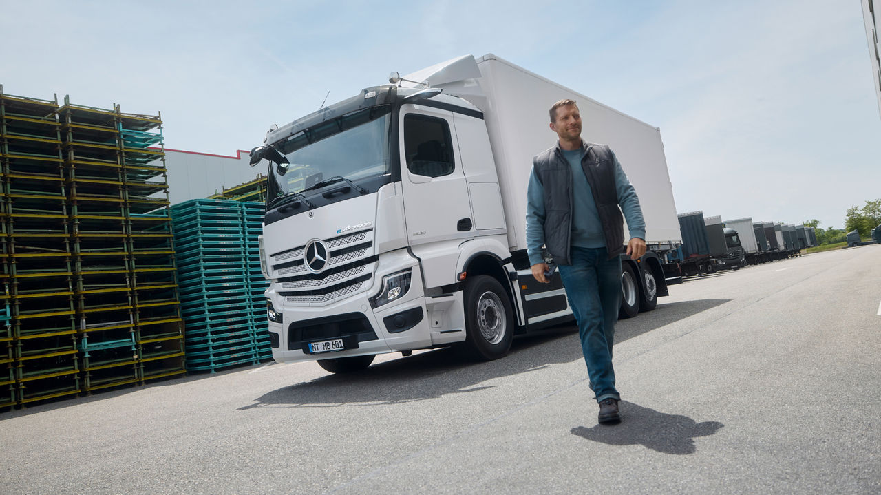 Een man in werkkleding loopt over een geasfalteerd oppervlak, terwijl op de achtergrond een witte eActros 600 van Mercedes-Benz Trucks met een aanhanger geparkeerd staat. Links zijn gestapelde pallets te zien en rechts een groot gebouw dat wijst op een magazijn- of industriecomplex.