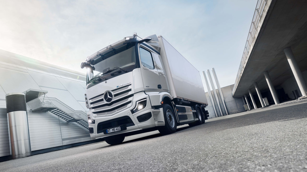 Een witte eActros 400 van Mercedes-Benz Trucks staat op een geasfalteerde ondergrond voor een modern gebouw met een buitentrap, waarbij de zon helder van linksboven schijnt. Rechts in beeld is een verhoogde betonconstructie met zuilen te zien, die een onderdoorgang of een overdekt gedeelte suggereert.