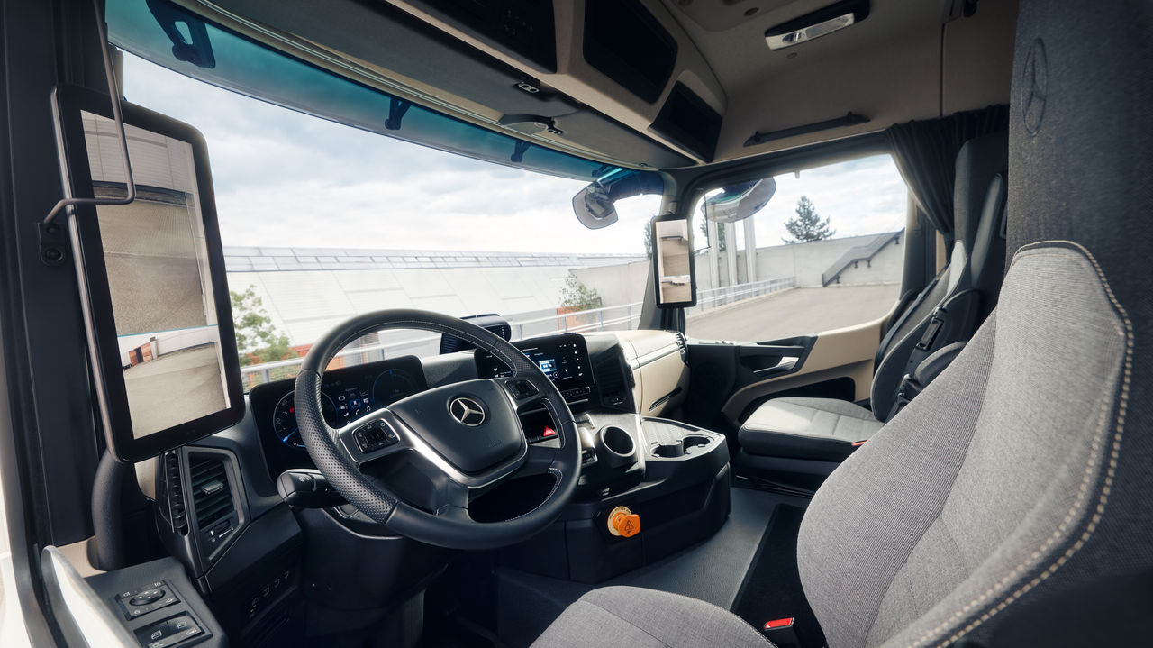 De afbeelding toont het interieur van de eActros van Mercedes‑Benz Trucks vanuit het perspectief van de passagiersstoel. Je ziet het stuur met de Mercedes‑ster, het dashboard met digitale displays en schermen, evenals de comfortabele stoelen in lichtgrijze stof.