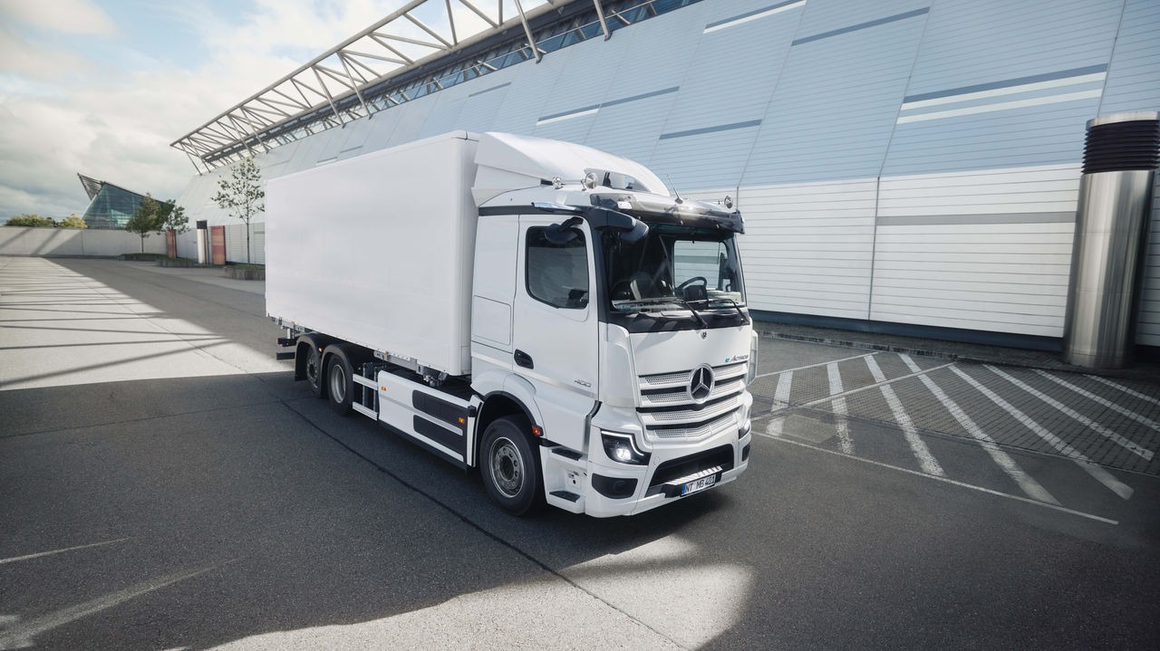 Een witte eActros 400 van Mercedes-Benz Trucks met een kofferopbouw staat op een geasfalteerde parkeerplaats voor een modern gebouw met een glazen gevel. Het zonlicht werpt lange schaduwen op de grond, wat duidt op een heldere dag.